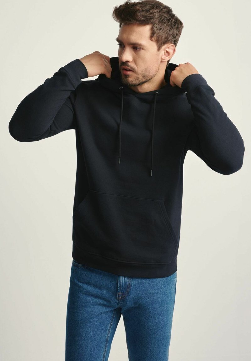 Mężczyzna poprawiający kaptur na czarnej bluzie z kapturem, ubrany w niebieskie dżinsy, na jednolitym tle.