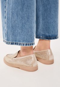 Beige Slippers mit strukturiertem, funkelndem Obermaterial und glatten Lederakzenten, mit einer hellbraunen Gummisohle. Getragen mit abgeknöpften blauen Jeans.