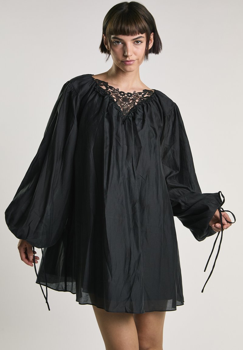 Junge Frau trägt ein locker sitzendes schwarzes Kleid mit Spitzenbesatz am Ausschnitt und langen, aufgeplusterten Ärmeln, die an den Handgelenken gebunden sind.