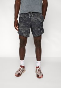 Man som bär svarta idrottsshorts med sprucken mönster, grå mönstrad T-shirt, vita strumpor och beige-röda löparskor står mot vit bakgrund.