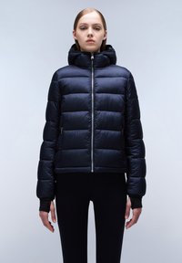 Marineblauwe pufferjack met kap, voorzien van een rits aan de voorkant, een gewatteerd patroon en elastische manchetten, gedragen over zwarte leggings. Gladde textuur.