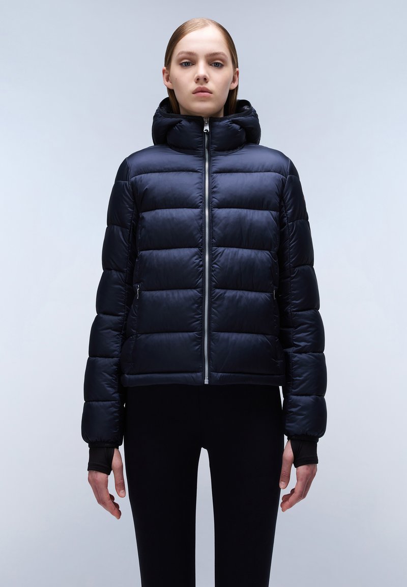 Marineblauwe pufferjack met kap, voorzien van een rits aan de voorkant, een gewatteerd patroon en elastische manchetten, gedragen over zwarte leggings. Gladde textuur.