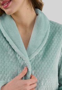 Mujer abrochándose un albornoz texturizado de color azul claro cerca del cuello, mostrando el suave tejido y el detalle del cuello.