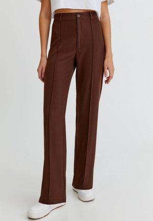 Frau trägt braune, hoch taillierte, gerade geschnittene Hose mit vorderem Nahtdetail und weißen Sneakers, steht vor einem schlichten Hintergrund.