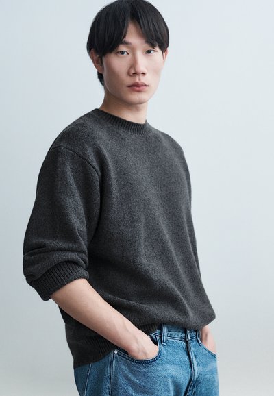 Un jeune homme aux cheveux noirs portant un pull gris foncé et un jean bleu, debout les mains dans les poches devant un fond clair uni.