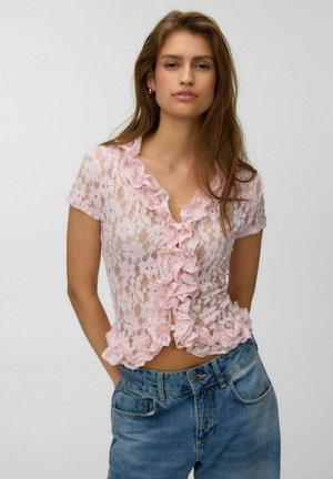 Jeune femme portant un chemisier court à manches courtes en dentelle florale rose avec des volants et un jean taille haute bleu, debout devant un fond uni.