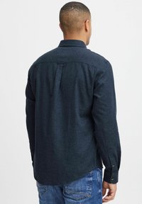 Temno modra dolgosleeve majica iz strukturirane tkanine, ki ima gumb na zapestju in zadnji pregib za dodatne podrobnosti.