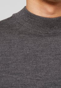 Nahaufnahme einer Person, die einen grauen gerippten Strickpullover mit Rollkragen trägt, wobei Details des Halses und des oberen Brustbereichs sichtbar sind.
