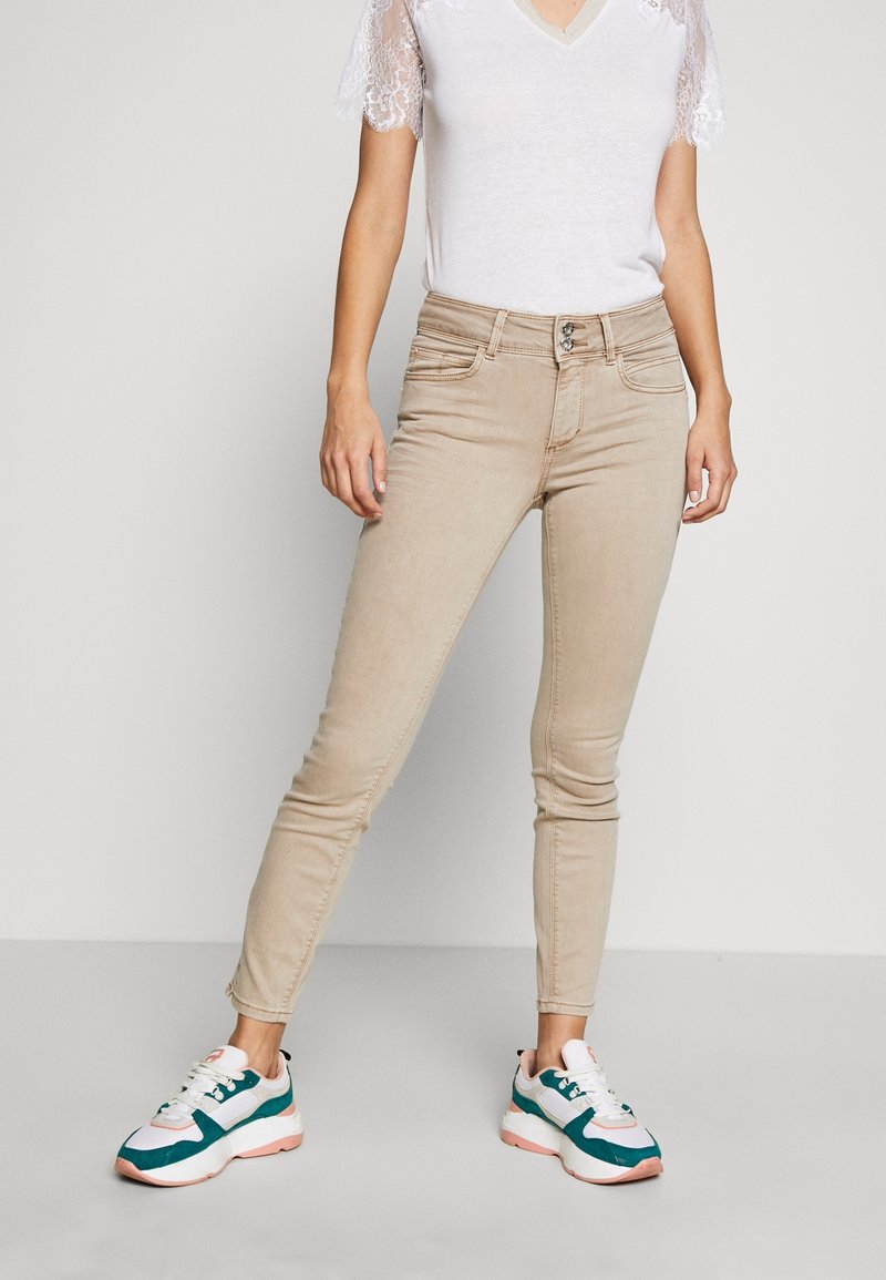Femme portant un jean skinny beige, un haut blanc à manches en dentelle, et des baskets blanches avec des détails verts et roses, debout devant un fond uni.