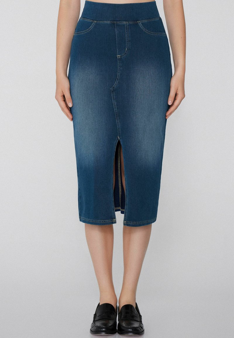 Mujer que lleva una falda de mezclilla azul de cintura alta y hasta la rodilla con una abertura frontal y unos mocasines negros sin cordones, de pie contra un fondo liso.