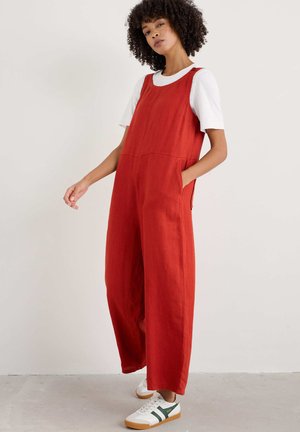 Rode linnen jumpsuit met een ontspannen pasvorm, wijde benen en zijzakken, gedragen over een wit T-shirt. Geaccentueerd met casual sneakers.