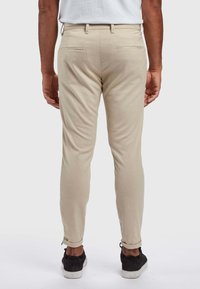 Mężczyzna ubrany w beżowe spodnie o dopasowanym kroju i czarne tenisówki, stojący tyłem na białym tle.