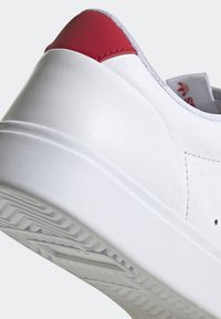 Vit sneaker med en röd hälaccent, slät texturmaterial, subtilt mönster på sidan och en ribbad vit sula.