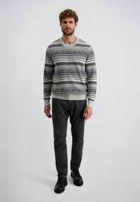 Brede gebreide trui in lichtgrijs met horizontale gestreepte patronen in donkerblauw, groen en crème. Gecombineerd met donkergrijze jeans en zwarte schoenen.