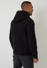 Sort strikket hoodie med ribbet tekstur, der har en stor hætte og lange ærmer. Båret med mørkegrå jeans, hvilket giver et afslappet look.
