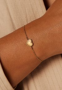 Goldene Kettenarmband mit einem kleinen herzförmigen Anhänger mit glatter Oberfläche und perlenbesetztem Rand, vor einer strukturierten Haut.