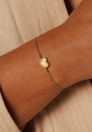Gouden kettingarmband met een kleine hartvormige bedel met een gladde oppervlakte en een geribbelde rand, geplaatst tegen een textuur van huid.