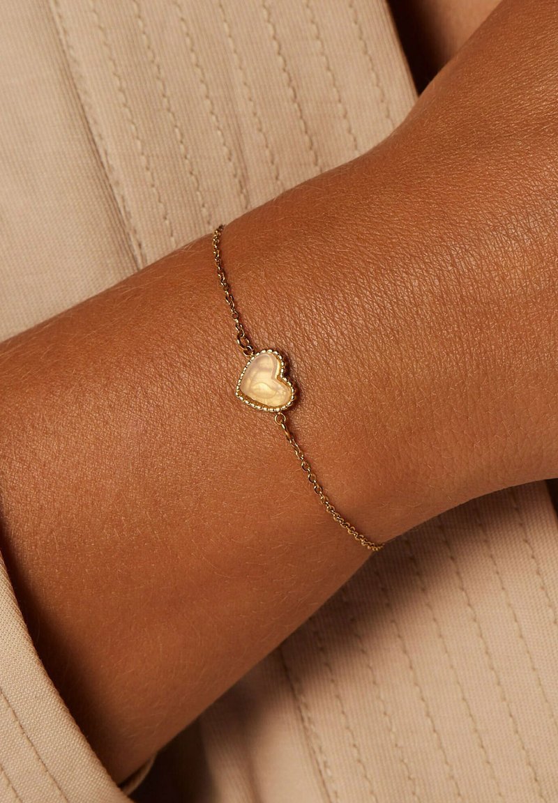 Goldene Kettenarmband mit einem kleinen herzförmigen Anhänger mit glatter Oberfläche und perlenbesetztem Rand, vor einer strukturierten Haut.