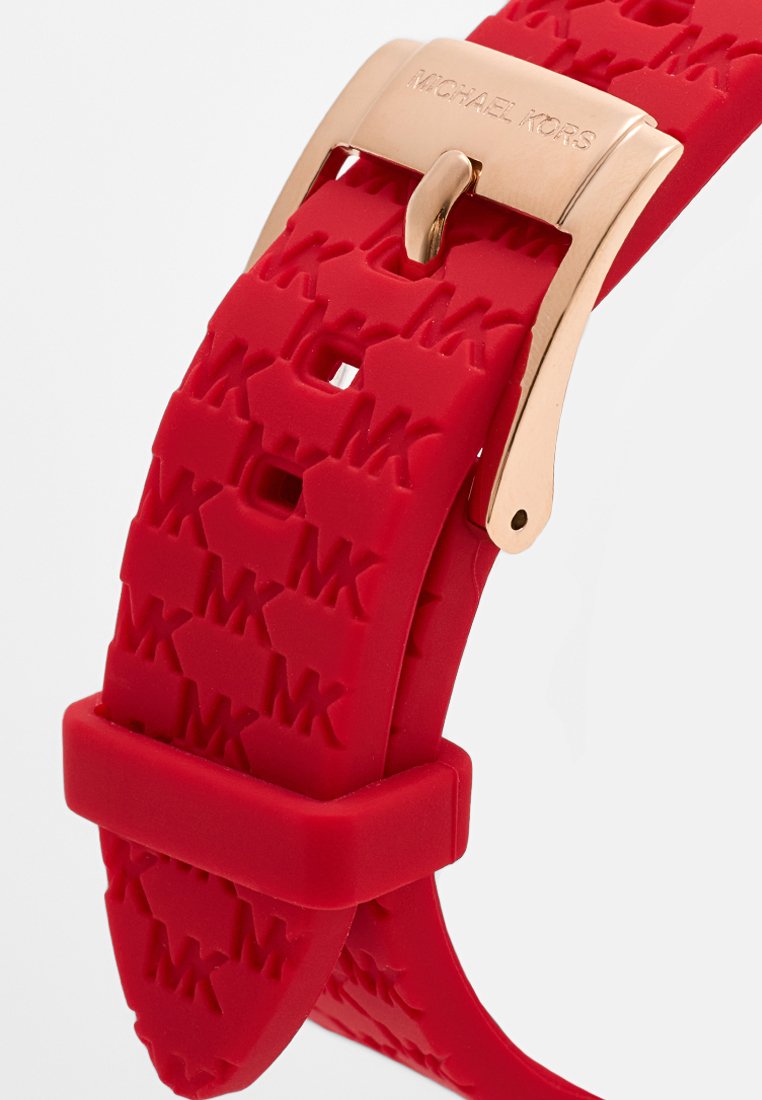 Bracelet de montre en silicone rouge avec un motif "MK" en relief, présentant une boucle en métal doré et une texture lisse et flexible.