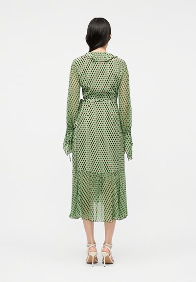 Γυναίκα που στέκεται με την πλάτη, φοράει πράσινο midi φόρεμα με μακριά μανίκια και μαύρες βούλες, καθώς και λευκά σανδάλια με ψηλό τακούνι.