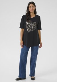 Camiseta negra de manga corta con un diseño de hoja metálica en el centro. Combinada con jeans de pierna ancha azul y bailarinas negras.