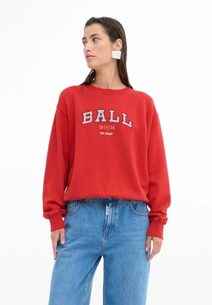 Rød sweatshirt med "BALL 1974 THE JEANS" trykt i hvid og blå skrift. Bæres med lyseblå jeans, der har en afslappet pasform.