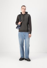 Hooded dark olive sweatshirt, light blue jeans, black shoes with textured design, holding a small blue bag with a patterned surface.