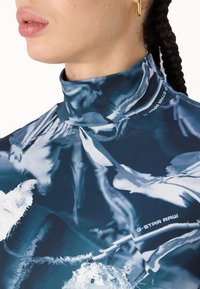 Μπλούζα κοντό μανίκι με ψηλό γιακά σε σκούρο μπλε χρώμα με αφηρημένα λευκά σχέδια. Φτιαγμένη από ομαλό, ελαστικό υλικό. Διαθέτει λογότυπο κοντά στον λαιμό.