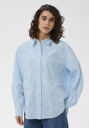 Femme aux cheveux bouclés portant une chemise à manches longues bleu clair à boutons avec des motifs de feuilles blanches, debout devant un fond uni clair.