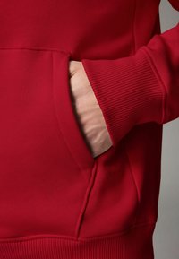 Felpa rossa con tasca a marsupio, polsini a coste e una consistenza morbida. Il materiale sembra spesso e confortevole, con cuciture pulite.