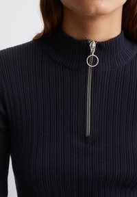 Frau trägt einen dunkelblauen gerippten Pullover mit einem silbernen Metallreißverschluss und einem runden Zugknopf am hohen Kragen.