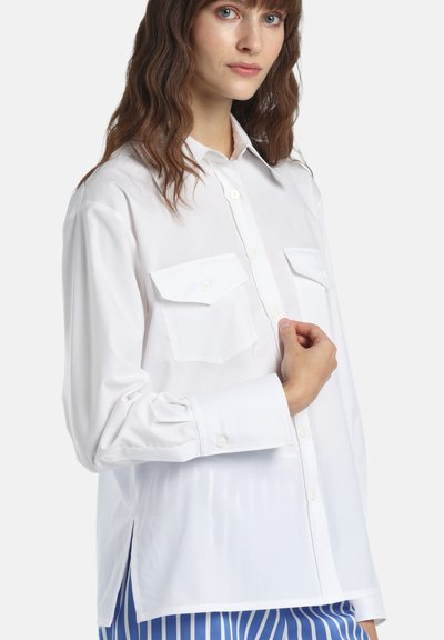 Femme aux cheveux bruns ondulés portant une chemise blanche à boutons avec des poches sur la poitrine, tenant un côté près du col, sur un fond uni.