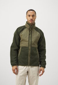 Jachetă de fleece verde, cu un panou frontal și un guler dintr-un material neted contrastant, fermoar complet și material texturat în întregime.