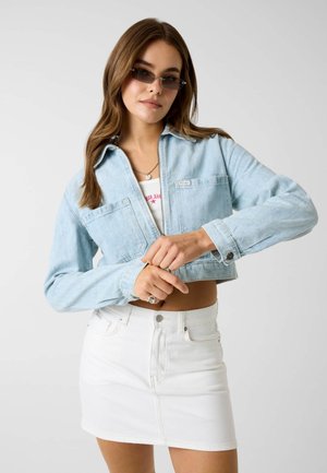 Kobieta w jasnoniebieskiej krótkiej kurtce dżinsowej, białym topie, białej mini spódniczce i małych prostokątnych okularach przeciwsłonecznych na jednolitym tle.