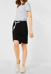 Femme portant un t-shirt rayé bleu clair et blanc, une jupe noire à cordon et des baskets blanches, se tenant devant un arrière-plan clair.