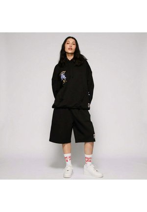 Giovane donna con felpa nera oversize, pantaloncini larghi neri, sneakers bianche e calzini bianchi con testo rosso su sfondo semplice.