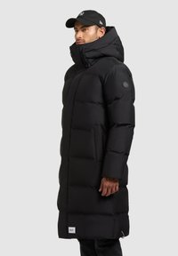 Sort pufferjakke med høj krave, quiltede syninger og et logo på ærmet. Har sidelommer og et afslappet design.