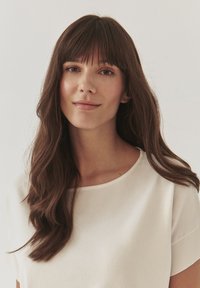 Portrait of a woman with long, wavy brown hair and blunt bangs, wearing a soft white shirt with a round neckline and short sleeves.
