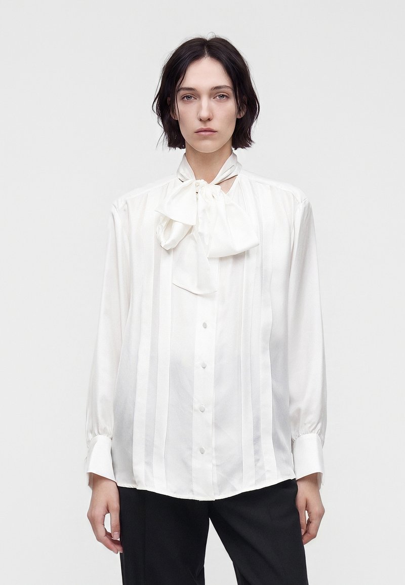 Femme aux cheveux courts noirs porte un chemisier blanc à manches longues avec des rayures verticales et un grand nœud noué au cou, associé à un pantalon noir.
