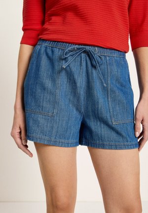 Frau trägt blaue Jeansshorts mit Kordelzug am Bund und großen Fronttaschen, kombiniert mit einem roten, gerippten Langarmshirt.