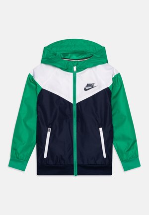 Chaqueta cortaviento de Nike con un diseño de bloques de color en verde, blanco y marino, cierre completo, capucha y bolsillos frontales con cremallera. Textura de tela suave.