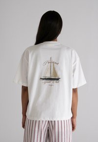 White cotton t-shirt featuring a sailboat graphic on the back with "Prohibited Yacht Club" text, paired with striped light pants.