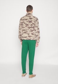 Fleece-pullover med camouflage-mönster i beige och brunt, matchad med gröna sweatpants och beige sneakers, visad bakifrån.