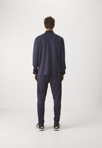 Marineblauwe trainingspak gemaakt van een gladde, lichtgewicht stof; heeft een langemouwen top en taps toegesneden broek met een elastische tailleband. Zwarte schoenen zichtbaar.