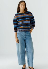 Bunt gestrickter Pullover mit blauen, orangefarbenen und schwarzen Streifen, kombiniert mit hellblauen, weit geschnittenen Jeans. Stricktextur sichtbar.