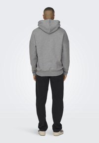 Sudadera gris con capucha, de corte relajado y hombros caídos, combinada con pantalones negros y zapatillas de color claro.