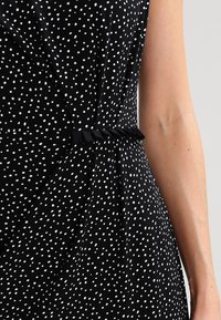 Black dress with white polka dots, featuring a pleated design and fabric belt. Smooth texture, sleeveless cut, accentuating a fitted waist.