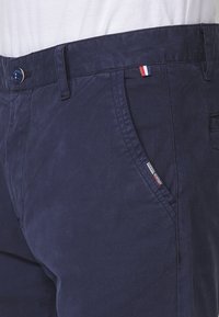 Pantalon bleu marine avec poches avant, taille boutonnée, passants de ceinture, petite étiquette tricolore rouge, blanc et bleu, porté avec une chemise blanche.