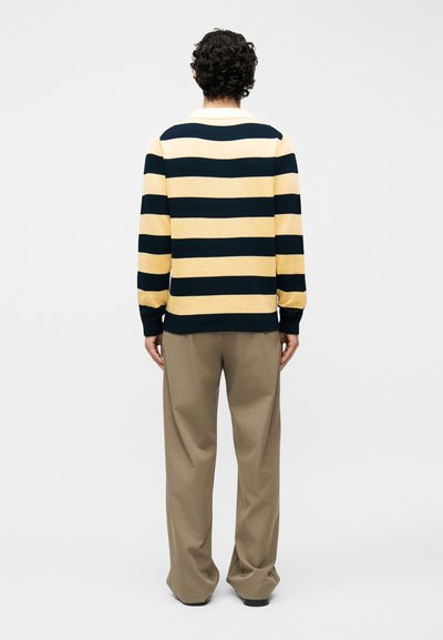 Homme aux cheveux bouclés portant une chemise à manches longues rayée horizontalement jaune et noire, ainsi qu'un pantalon beige ample, debout, vue de dos.