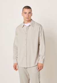 Mladić s kratkom kosom, obučen u bež manžestrovanu košulju na dugmad i odgovarajuće hlače, stoji nasuprot jednobojnoj svijetloj pozadini.